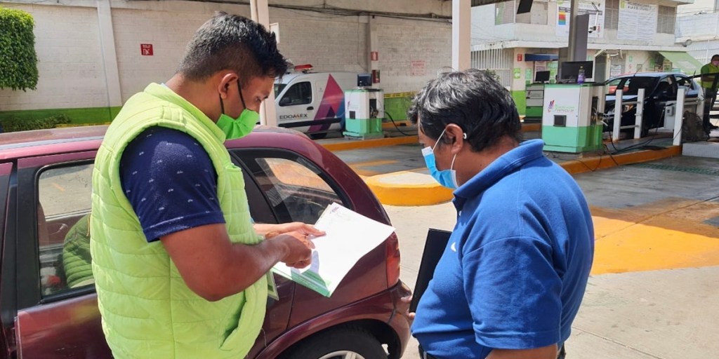 Dos personas en un verificentro revisando documentos de un vehículo. Uno de ellos lleva un chaleco verde y el otro una camisa azul. Se observan máquinas de verificación al fondo.