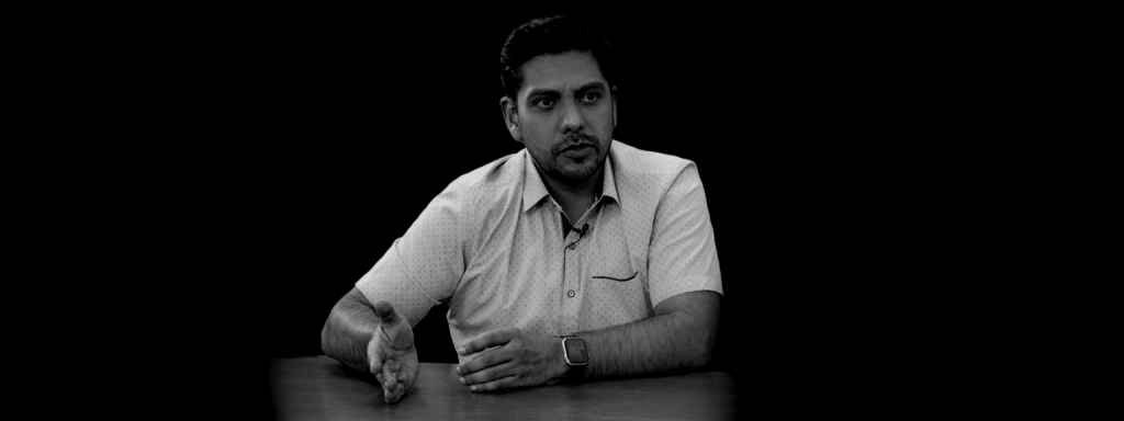 Hombre sentado en una mesa, hablando y gesticulando, con fondo negro.