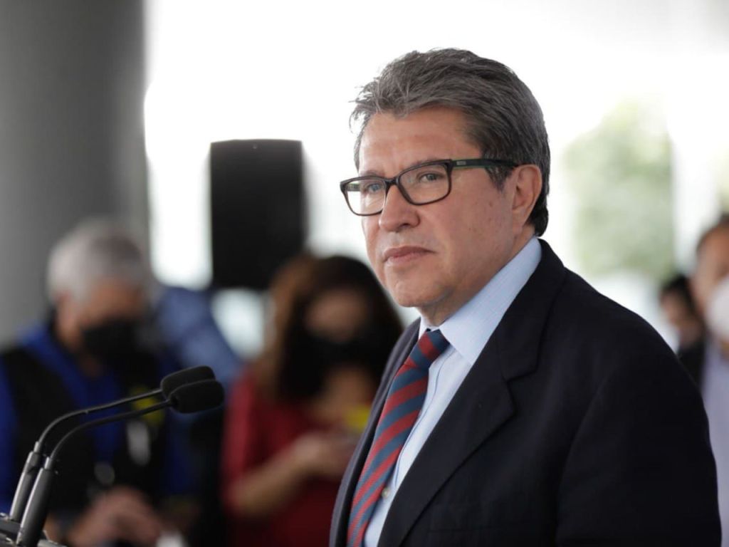 Retrato de un funcionario en un evento público, con un micrófono frente a él y un fondo borroso de asistentes.