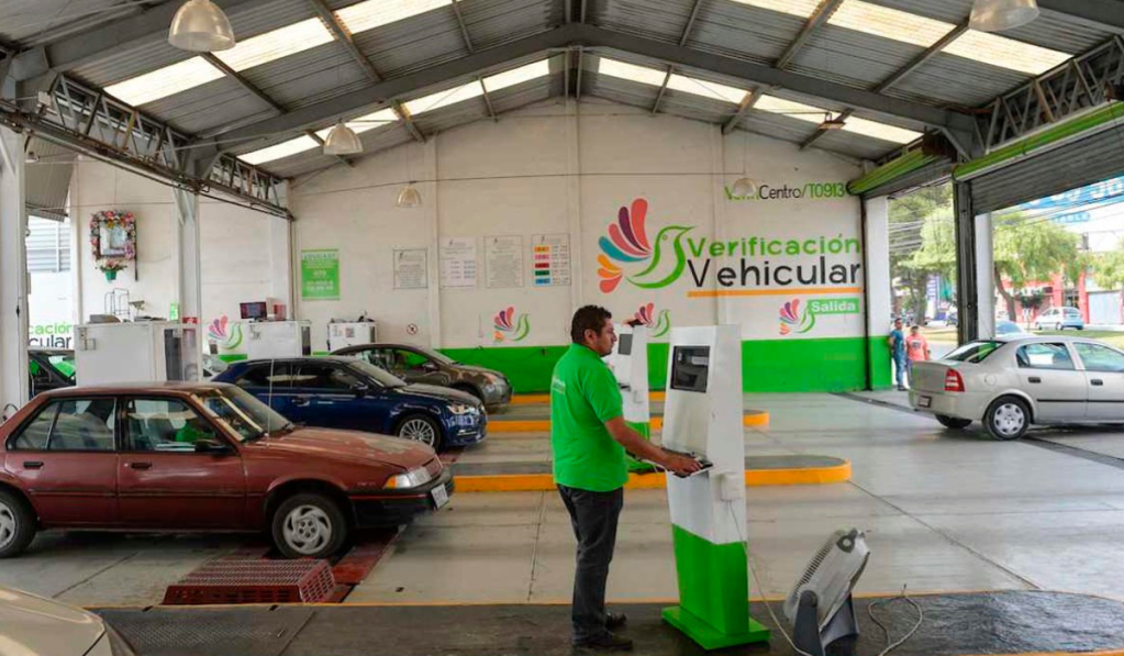 Centro de verificación vehicular con varios autos en espera y un empleado atendiendo un kiosco de verificación.