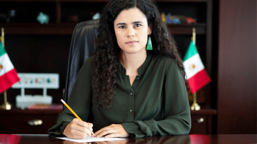 Mujer sentada en un escritorio, escribiendo con un lápiz mientras mira a la cámara. Detrás hay banderas de México.