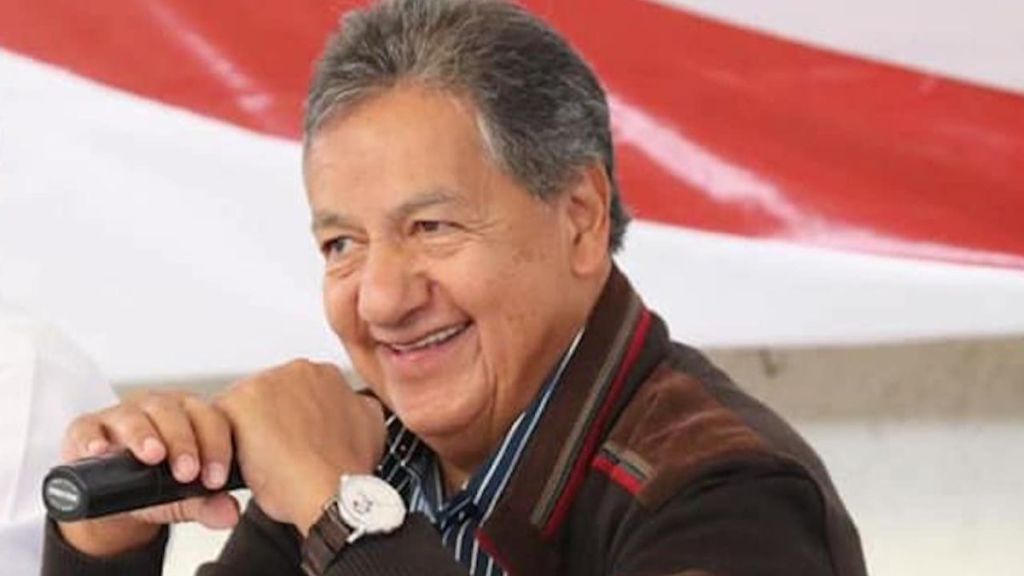 Hombre sonriendo mientras sostiene un micrófono en una mesa, con fondo de tela blanca y roja.