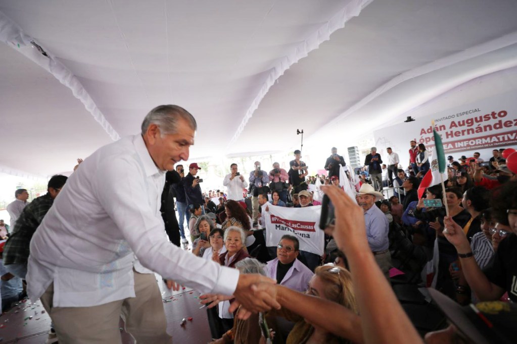 A political figure shakes hands with a supporter during a public event, surrounded by an engaged audience holding banners and flags.