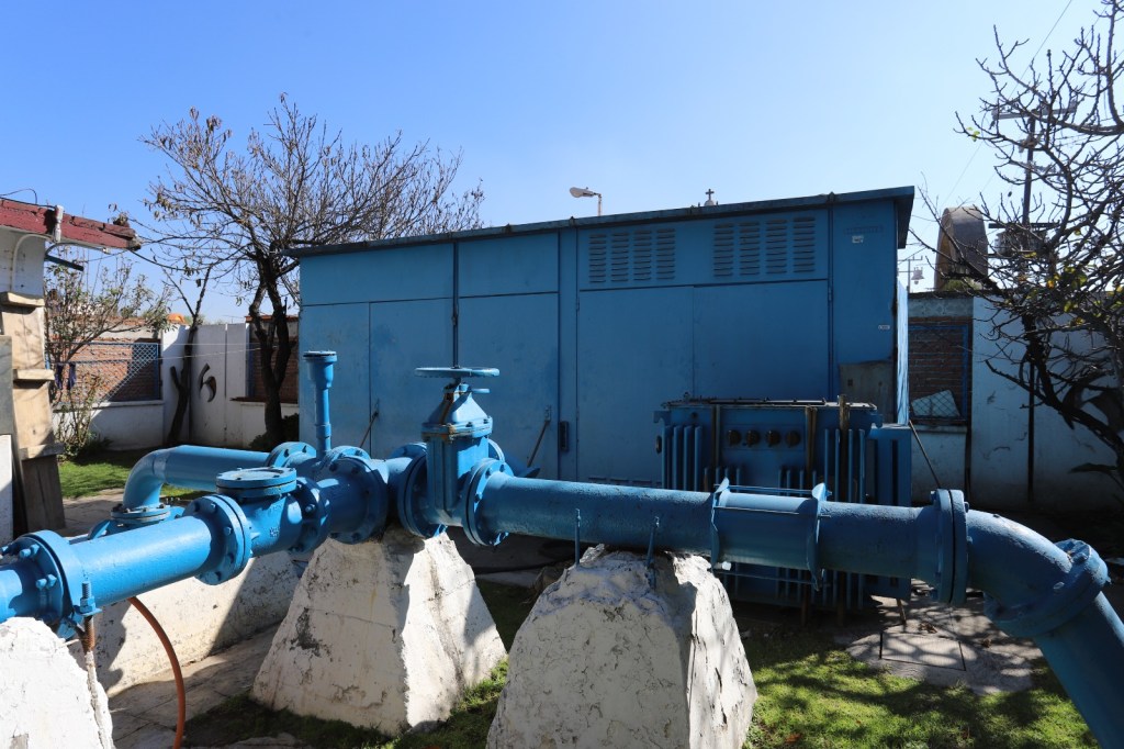A blue water pump system with large pipes situated in a garden area, next to a blue utility building and trees against a clear sky.