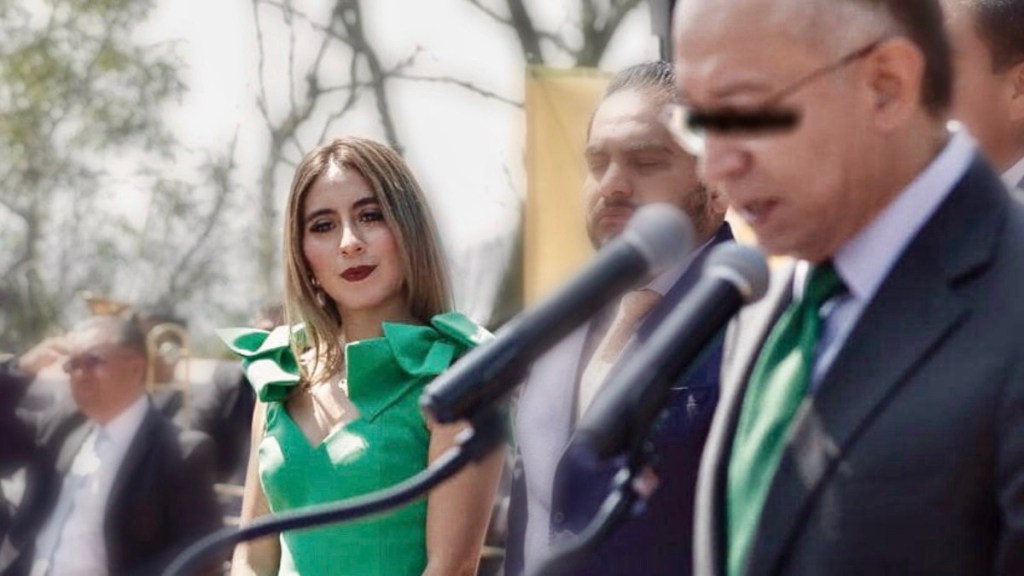 Una mujer con vestido verde, asistiendo a un evento junto a otros están en el fondo, donde un hombre habla en un micrófono.