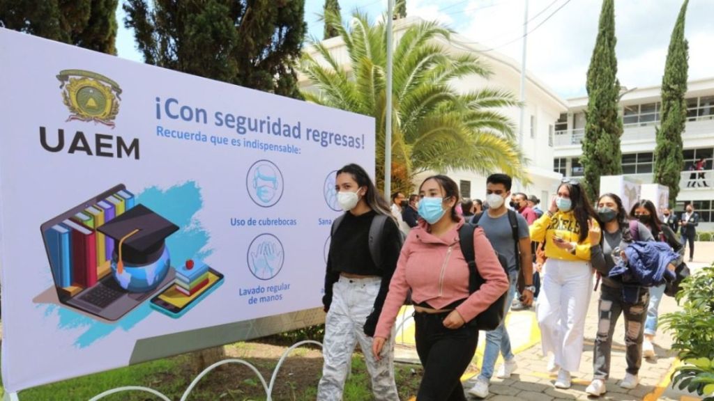Students walking past a sign with health guidelines for returning to campus at UAEM, including mask usage and hand washing.