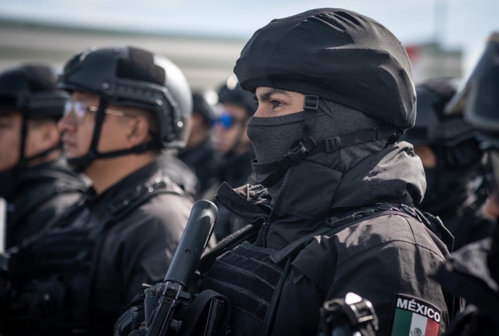 Grupo de oficiales de policía vestidos con uniformes negros y equipos de protección, observando atentamente durante una formación.