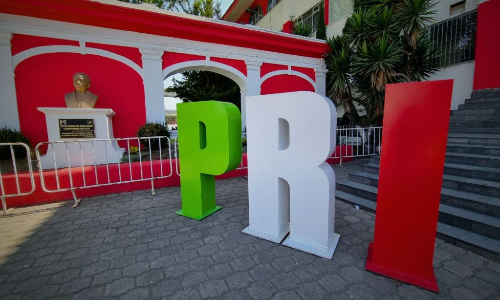 Letreros grandes de 'PRI' en colores verde, blanco y rojo frente a un busto en una fachada pintada de rojo.