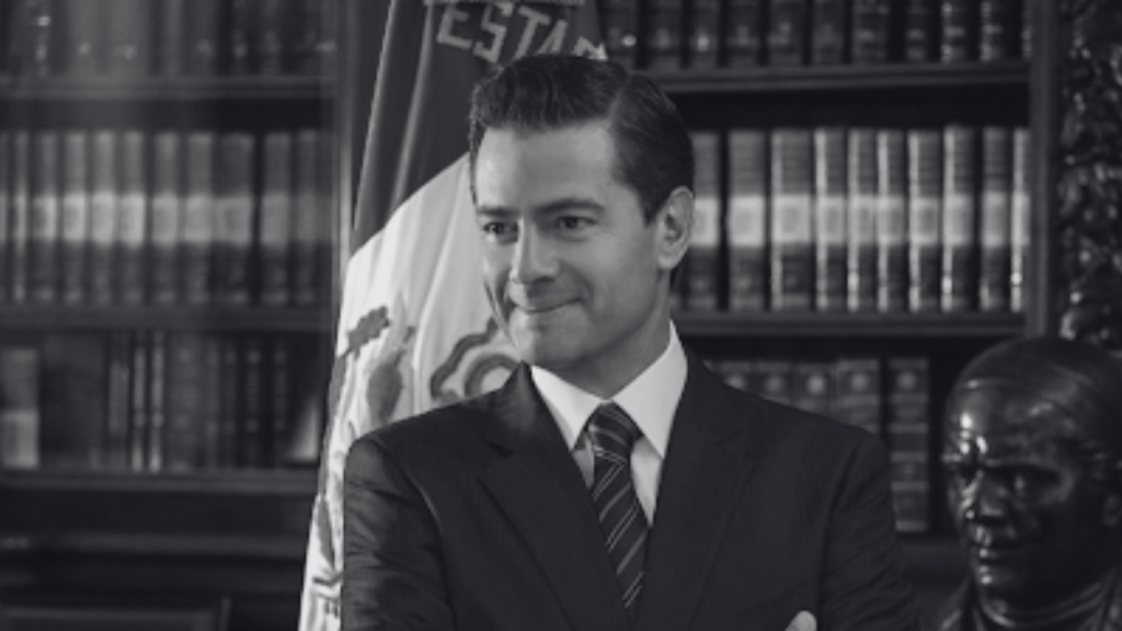 Hombre sonriendo en un entorno formal con una biblioteca y una bandera de México de fondo.