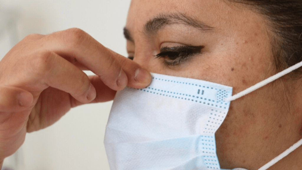 Close-up of a woman adjusting a blue medical face mask on her face.