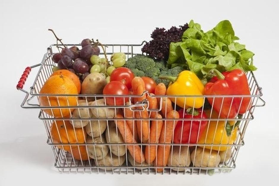 Cesta de supermercado llena de frutas y verduras variadas, incluyendo naranjas, uvas, brócoli, lechuga, pimientos y zanahorias.