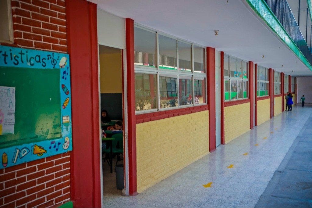 Pasillo de una escuela con aulas a ambos lados, decorado con un mural colorido. Se pueden ver estudiantes en el interior, mientras que otros caminan por el pasillo.