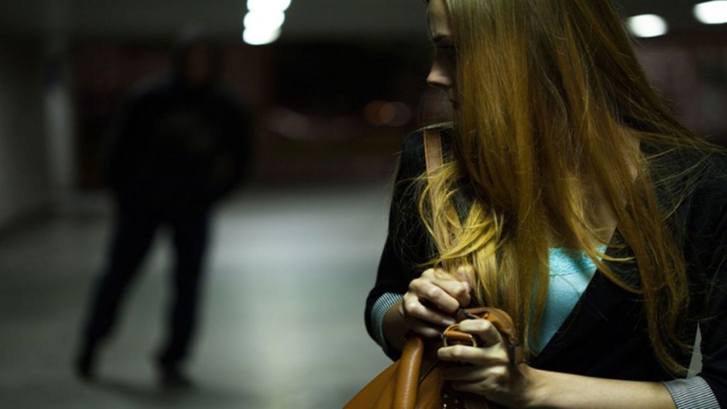 Una mujer con cabello largo y rubio se muestra asustada en un lugar oscuro, mientras un hombre en la distancia parece estar acercándose.