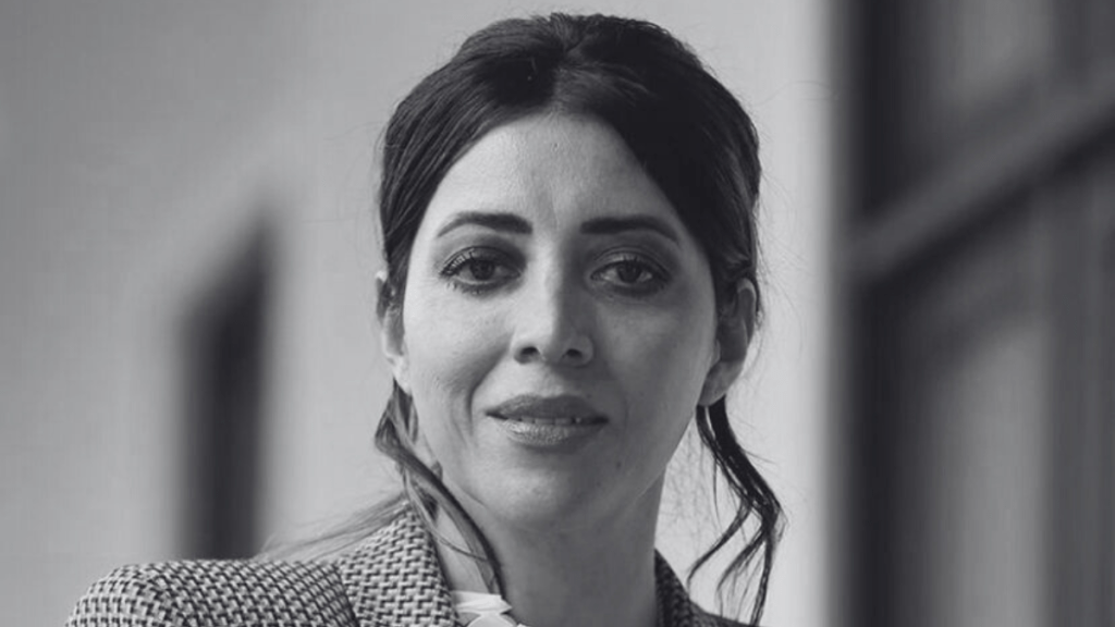 Close-up portrait of a woman with long dark hair, wearing a checked blazer and looking directly at the camera.