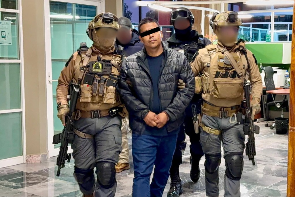 Un hombre es escoltado por dos agentes de seguridad, vestidos con equipo táctico, en un interior de oficina.