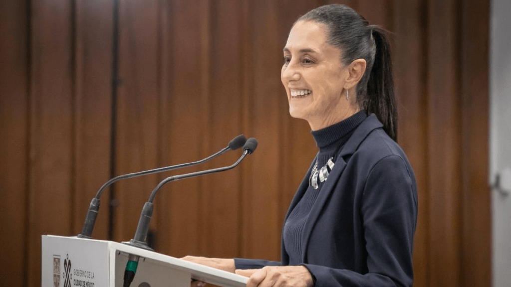 Mujer sonriente hablando en un podio, con cabello recogido y vestimenta formal en un entorno interior.
