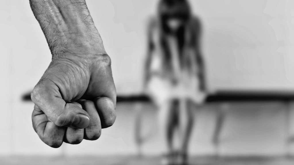 Un puño cerrado en primer plano, con una figura femenina en segundo plano que parece estar triste o asustada, enfatizando un tema de violencia y agresión.