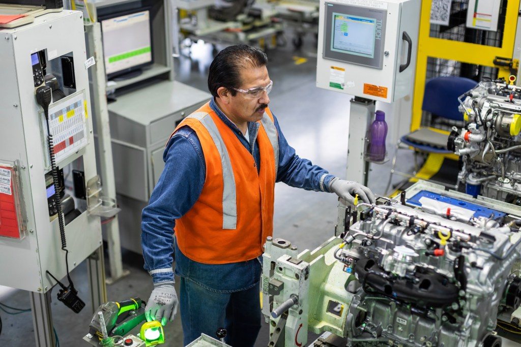 Un trabajador en una línea de ensamblaje industrial, luciendo un chaleco reflectante, inspecciona un motor en un entorno de fabricación.
