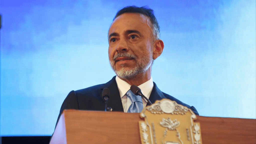 Hombre de negocios hablando en un podio, con una camisa blanca y una corbata azul, en un evento con fondo azul.