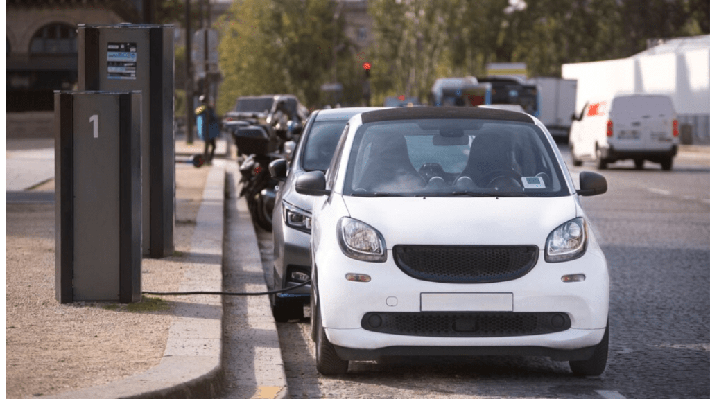 Coche eléctrico blanco enchufado a una estación de carga en una calle, con otros vehículos estacionados alrededor.