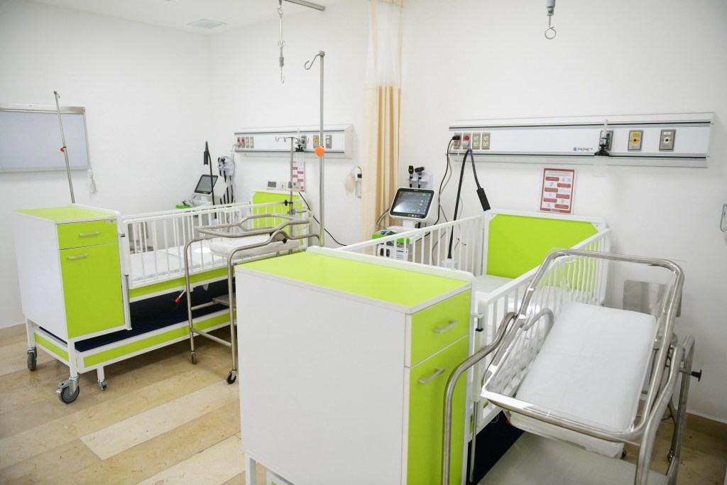 Sala de hospital con cunas y equipo médico, paredes blancas y decoración en tonos verdes.