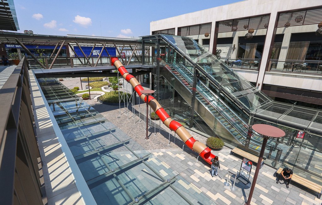 Vista aérea de un centro comercial con una resbaladilla roja y marrón, escalera mecánica y áreas ajardinadas.