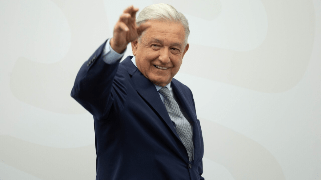 A smiling man in a suit waves with his right hand, against a light background.