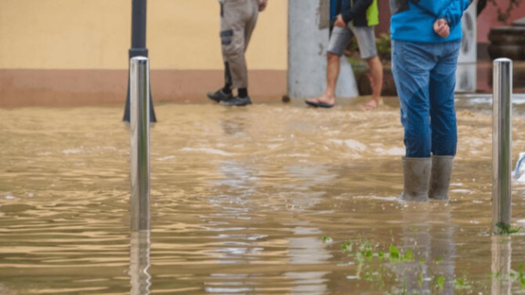 Personas caminando en una calle inundada con agua cubriendo el terreno y postes visibles.