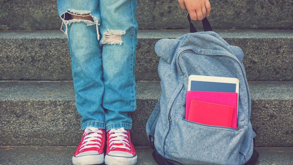 Un niño con pantalones rotos y zapatillas rojas sostiene una mochila gris con libros en los escalones.