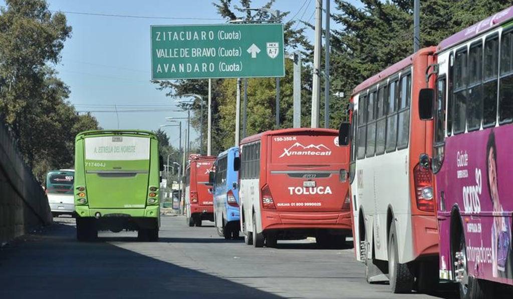 A view of several buses on a street with a road sign indicating directions to Zitácuaro, Valle de Bravo, and Avándaro.