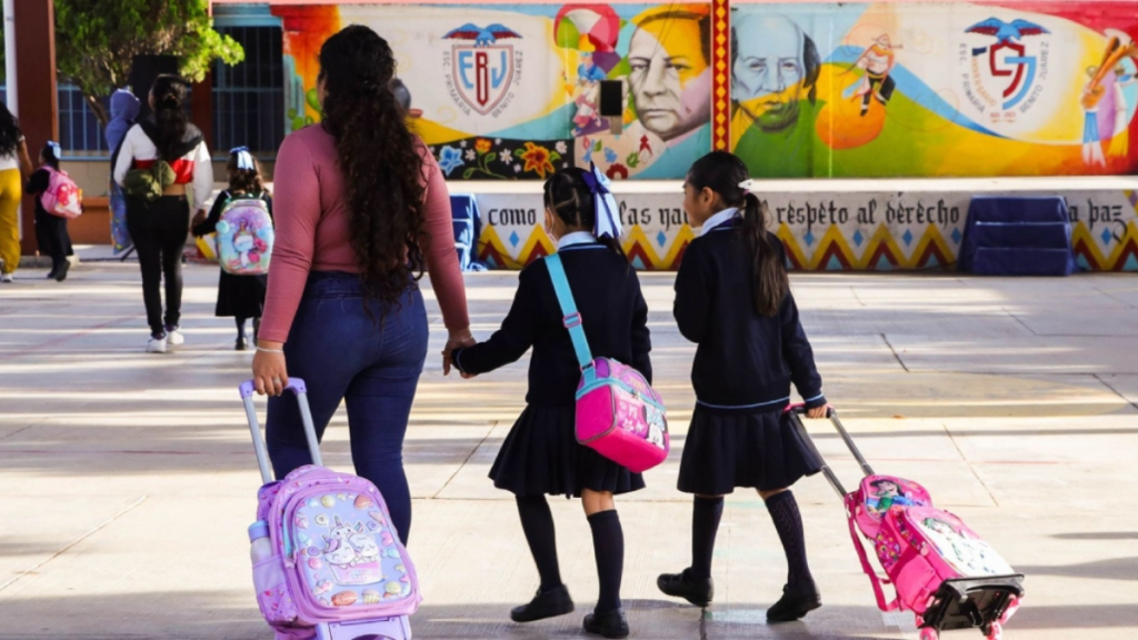 Una madre caminando con dos niñas hacia una escuela, cada una con mochilas coloridas. Al fondo, se ve un mural artístico.