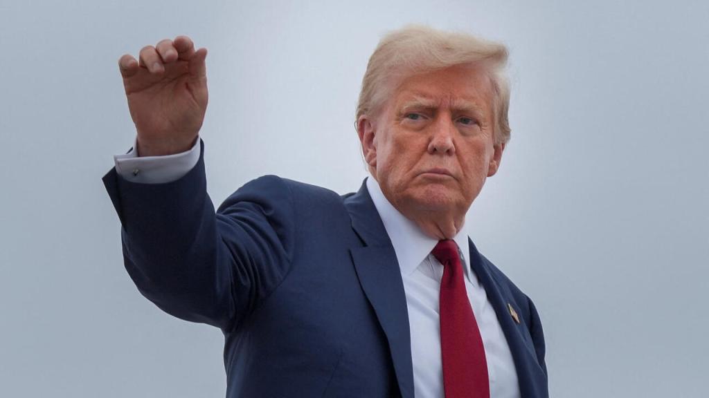 Hombre en traje oscuro con corbata roja, saludando con la mano levantada frente a un cielo nublado.