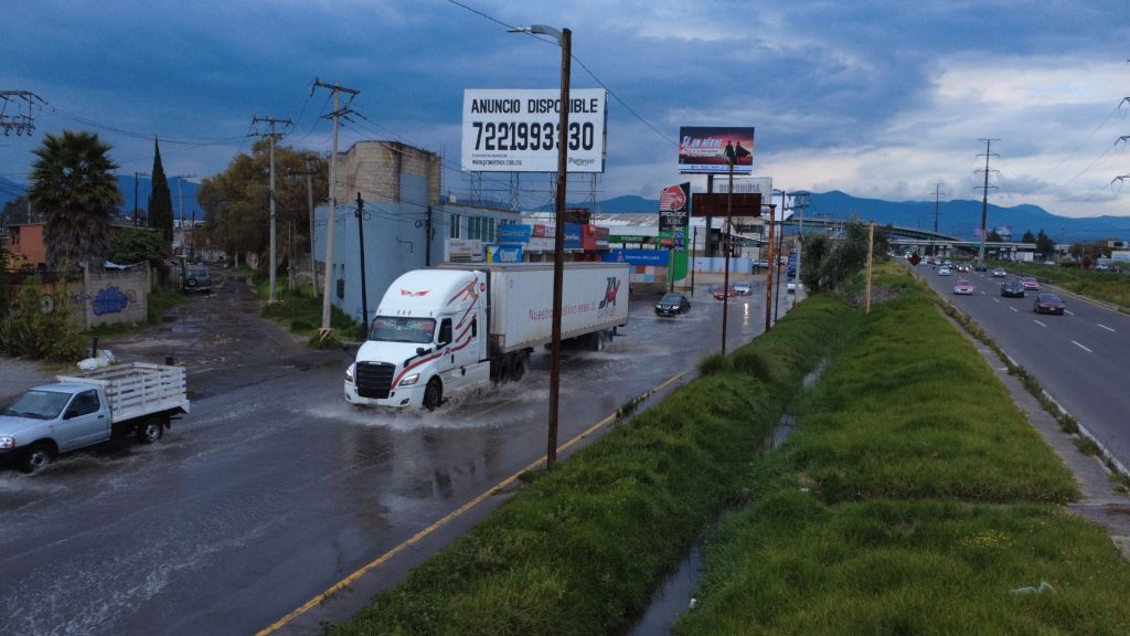 Una carretera inundada con vehículos, incluyendo un camión grande y una camioneta, bajo un cielo nublado. Se observan letreros publicitarios y montañas al fondo.