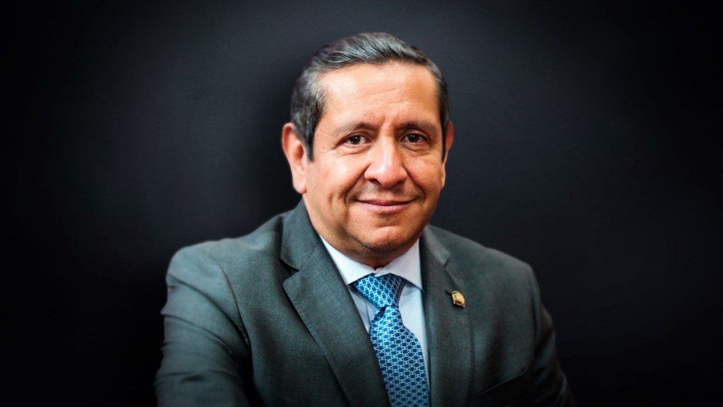 Retrato de un hombre sonriente con traje gris y corbata azul, frente a un fondo oscuro.