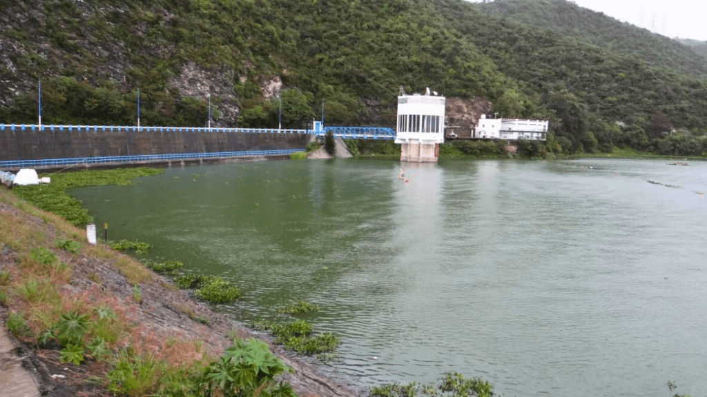 Vista de un embalse con agua verde y vegetación alrededor, acompañado por un edificio de control en la orilla.