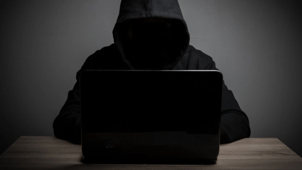 Persona en la sombra con capucha frente a un ordenador portátil en una mesa, en un entorno oscuro.
