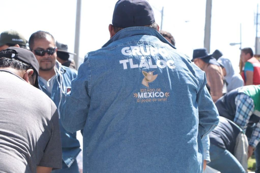 A group of people outdoors with one individual in a blue denim jacket featuring the text 'GRUPO TLALOC' and 'ESTADO DE MEXICO' on the back, engaged in an activity.