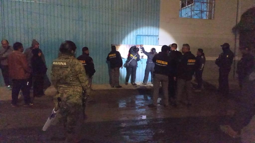 Grupo de personas y autoridades cerca de una puerta azul en un ambiente nocturno, algunos con chaquetas de la 'Fiscalía' y otros de la 'Marina'.