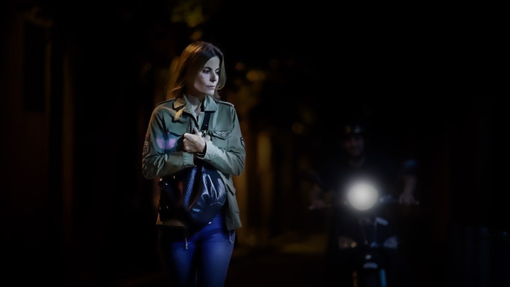 Una mujer caminando por una calle oscura, con una chaqueta y un bolso, mientras un motociclista avanza hacia ella con la luz encendida.