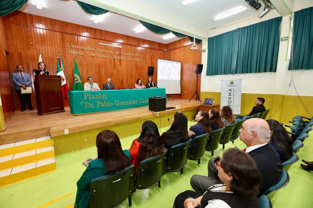 A conference or event taking place at the Universidad Autonoma del Estado de Mexico, featuring a speaker at a podium with a backdrop of the institution's name and a seated audience in front.