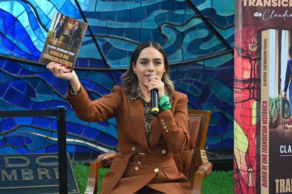 Claudia Sheinbaum, presidenta de México, sostiene un libro titulado 'Diario de una Transición Histórica' durante un evento, mientras habla a un micrófono en un ambiente decorado con mosaicos.