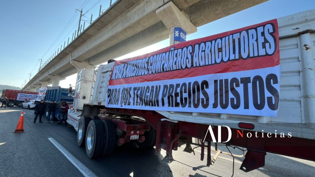 Camiones parados en la carretera con una pancarta que exige precios justos para los agricultores, bajo un puente elevado.