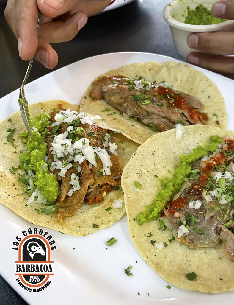 Tacos de barbacoa servidos en plato, con carne, cebolla picada, salsa verde y guacamole, mientras se sostiene un tenedor en una mano.