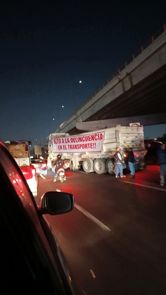 Un camión de carga bloquea la carretera con un cartel que dice 'ALTO A LAS DESERCIÓN EN EL TRANSPORTE', causando congestión vehicular durante la manifestación.