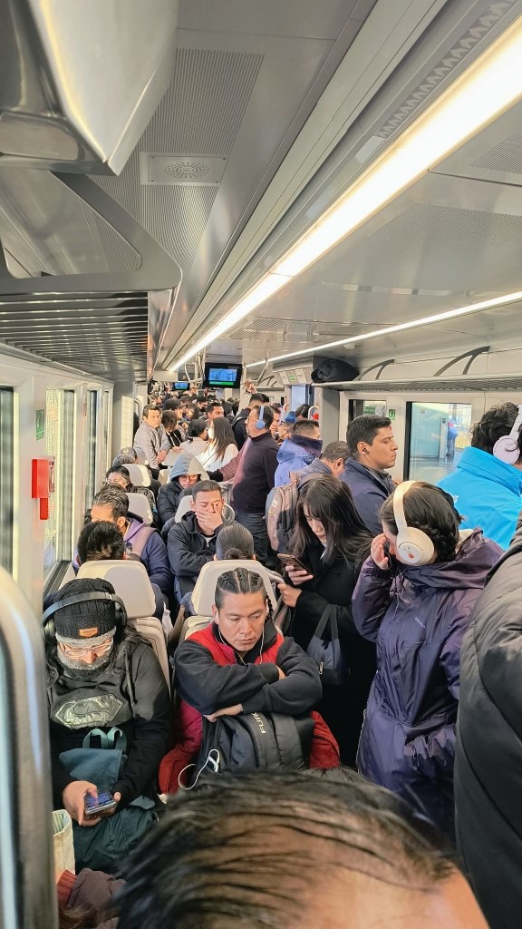 Interior de un tren interurbano abarrotado, con pasajeros de pie y sentados, algunos usando audífonos, en un ambiente de alta demanda.
