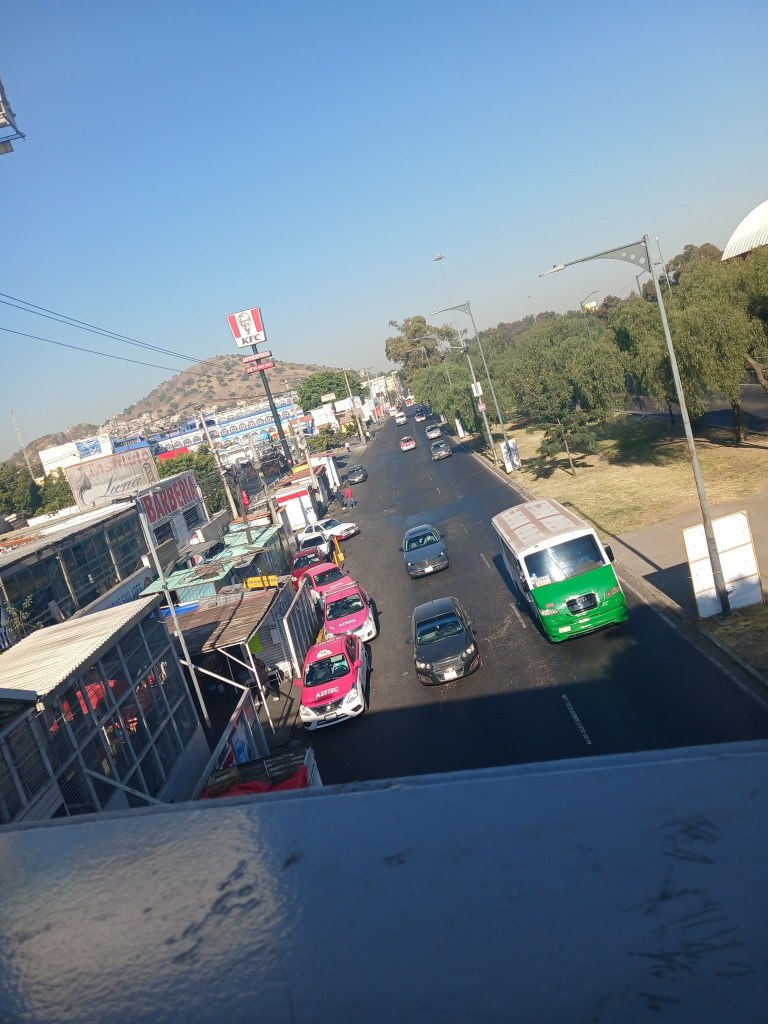 Vista de una calle en México con tráfico moderado, incluyendo taxis rosa y vehículos particulares, y un local de KFC visible en el fondo.