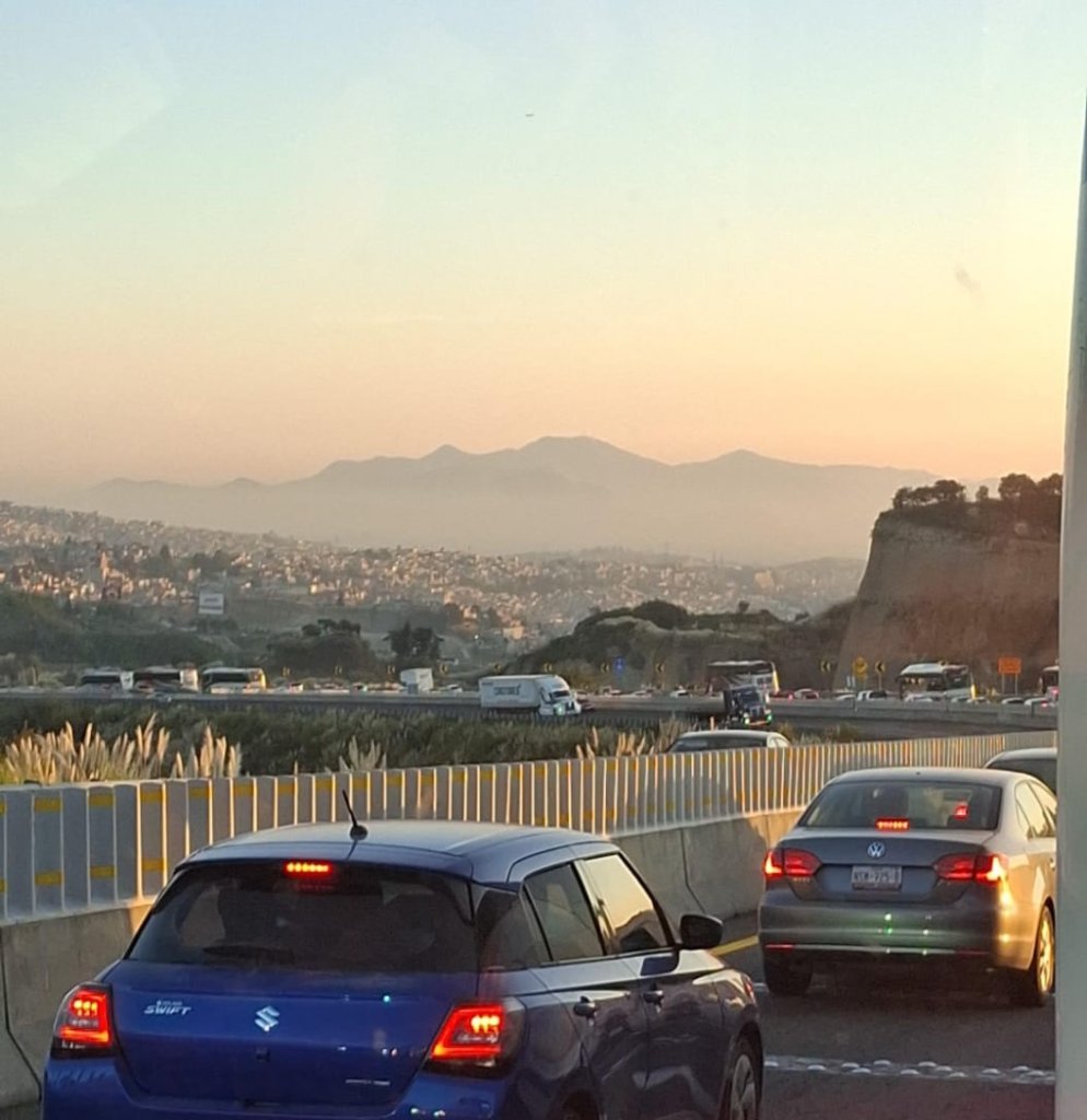 Vista de una carretera con tráfico a la hora del amanecer, mostrando vehículos en movimiento y montañas al fondo.