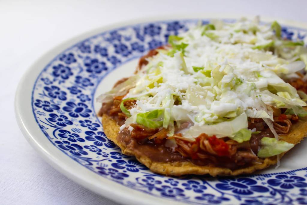 Un plato de sope con frijoles, carne deshebrada, lechuga y queso, servido en un plato azul decorado con flores.