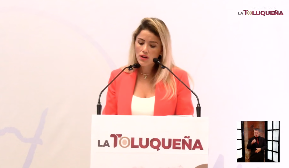 Una mujer hablando en un podio con un micrófono, promoviendo la Escuela Municipal de Emprendimiento en Toluca, con una pantalla al lado que muestra interpretación en lengua de señas.