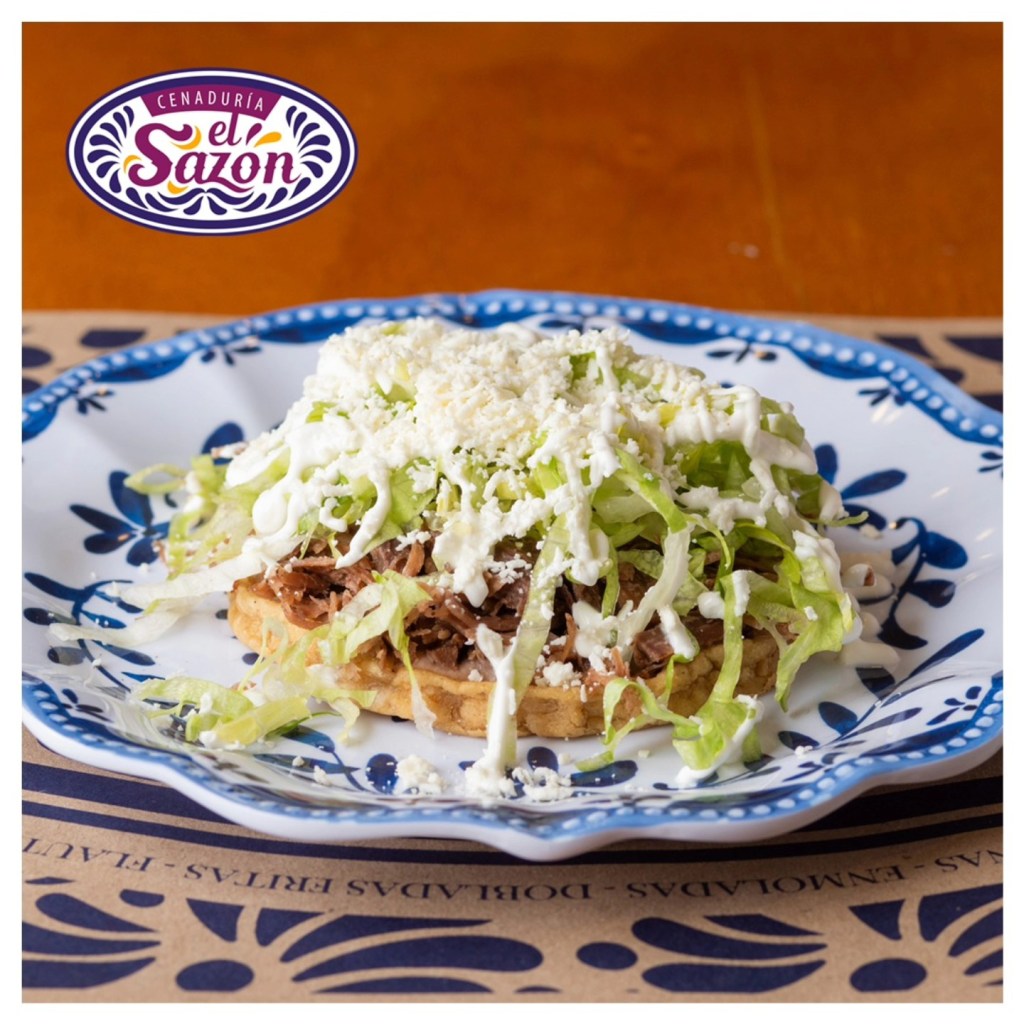 Un plato de sope con carne deshebrada, lechuga, crema y queso, servido en un plato azul decorativo. Logo de Cenaduría El Sazón en la parte superior.